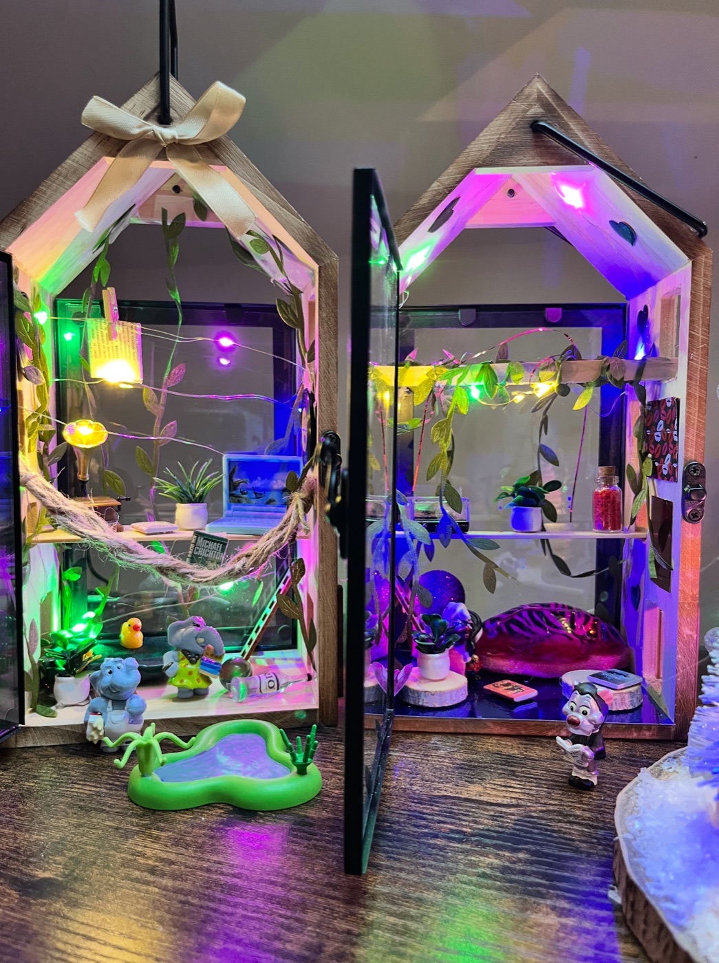 Two lit miniature glass houses with tiny elephants, plants, vines, and a pool